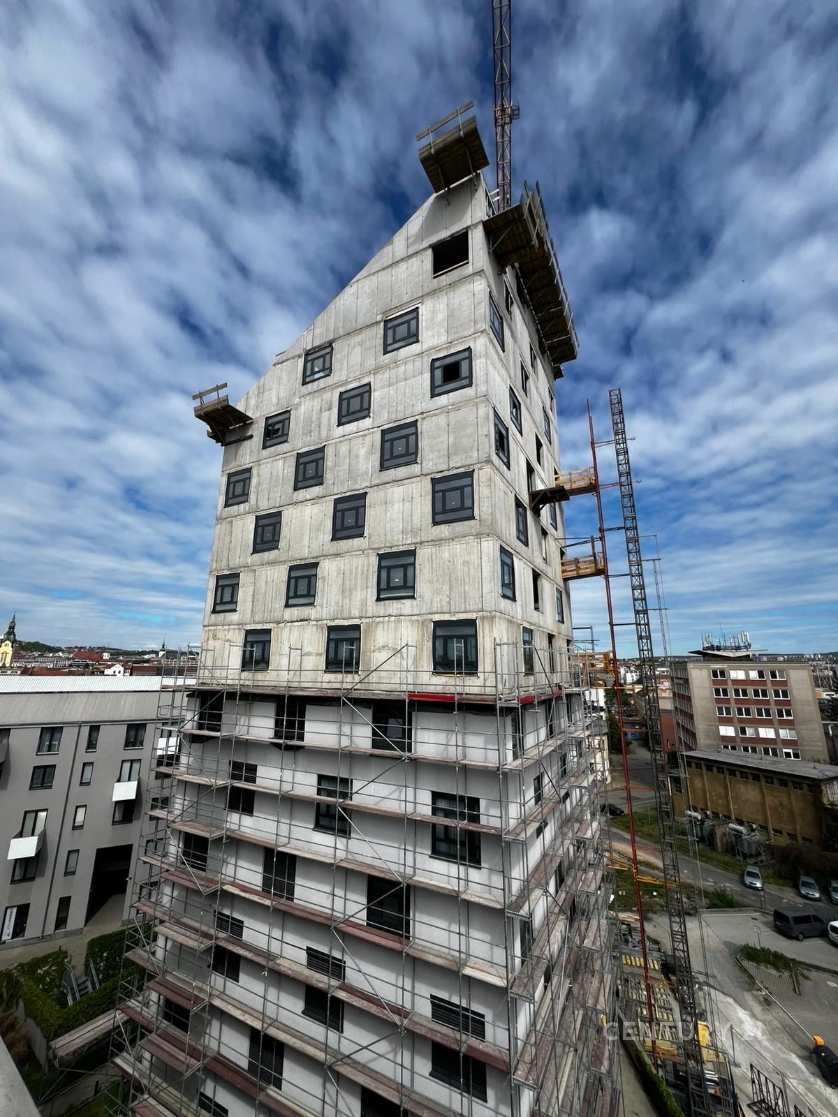 Nově budovaná bytová budova s moderním designem, betonovým povrchem a velkými okny, nacházející se v rozvíjející se městské lokalitě.