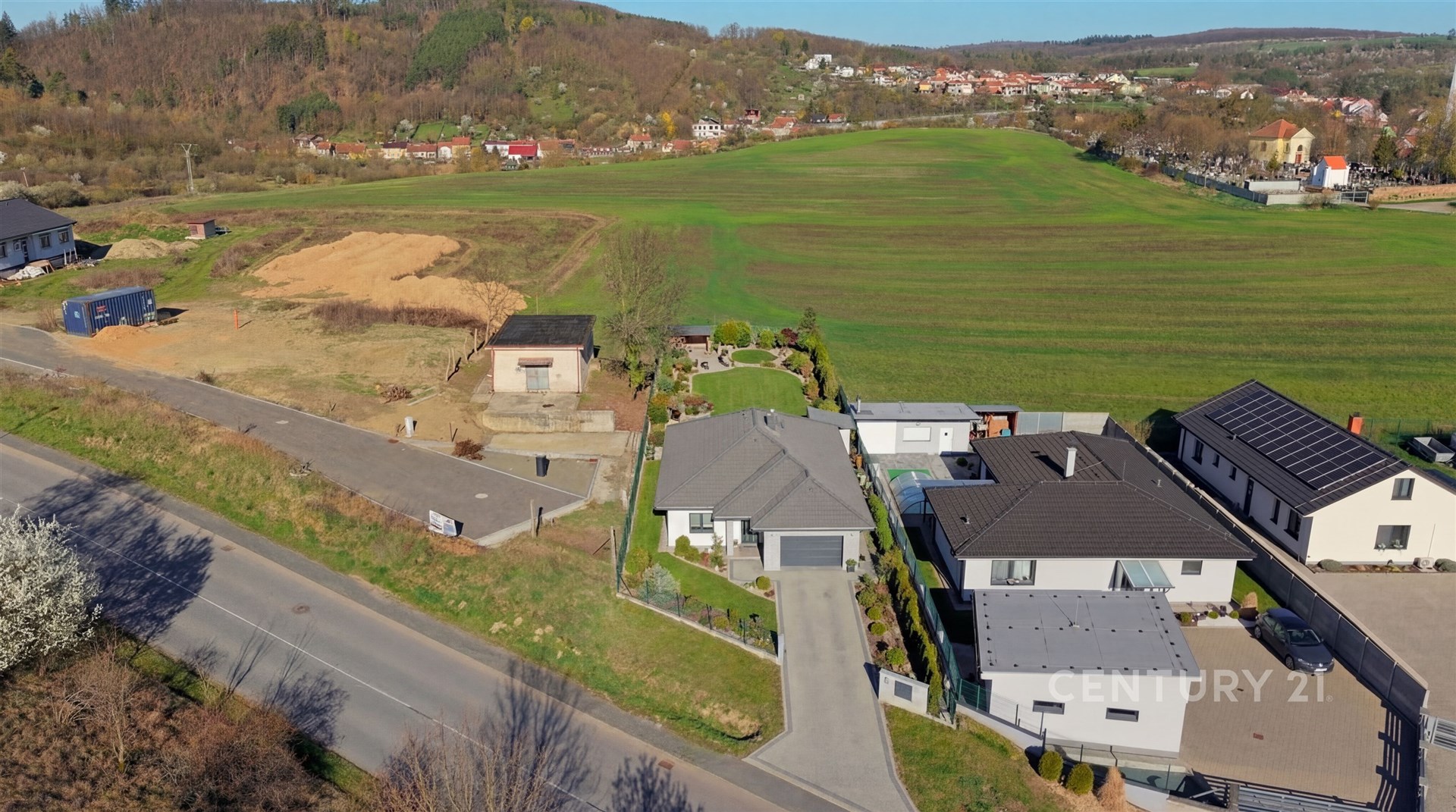 Pohled na moderní rodinný dům s velkými okny a zahradou, nacházející se v klidné oblasti s výhledem na louku a okolní krajinu.
