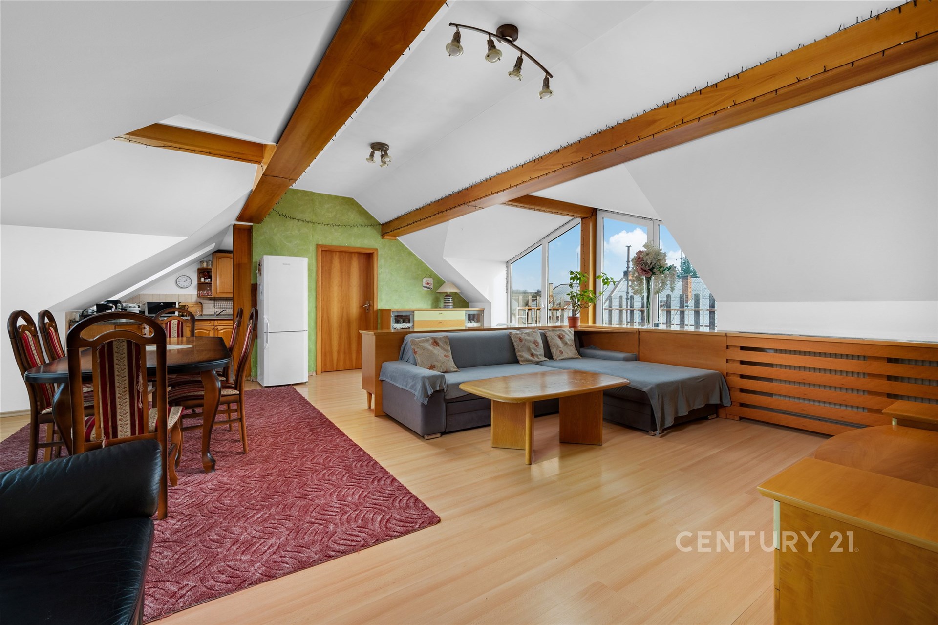 Podkrovní obývací pokoj s parketovou podlahou a velkými okny, moderní kuchyňský kout a dřevěné stropní trámy, světlý a prostorný prostor.