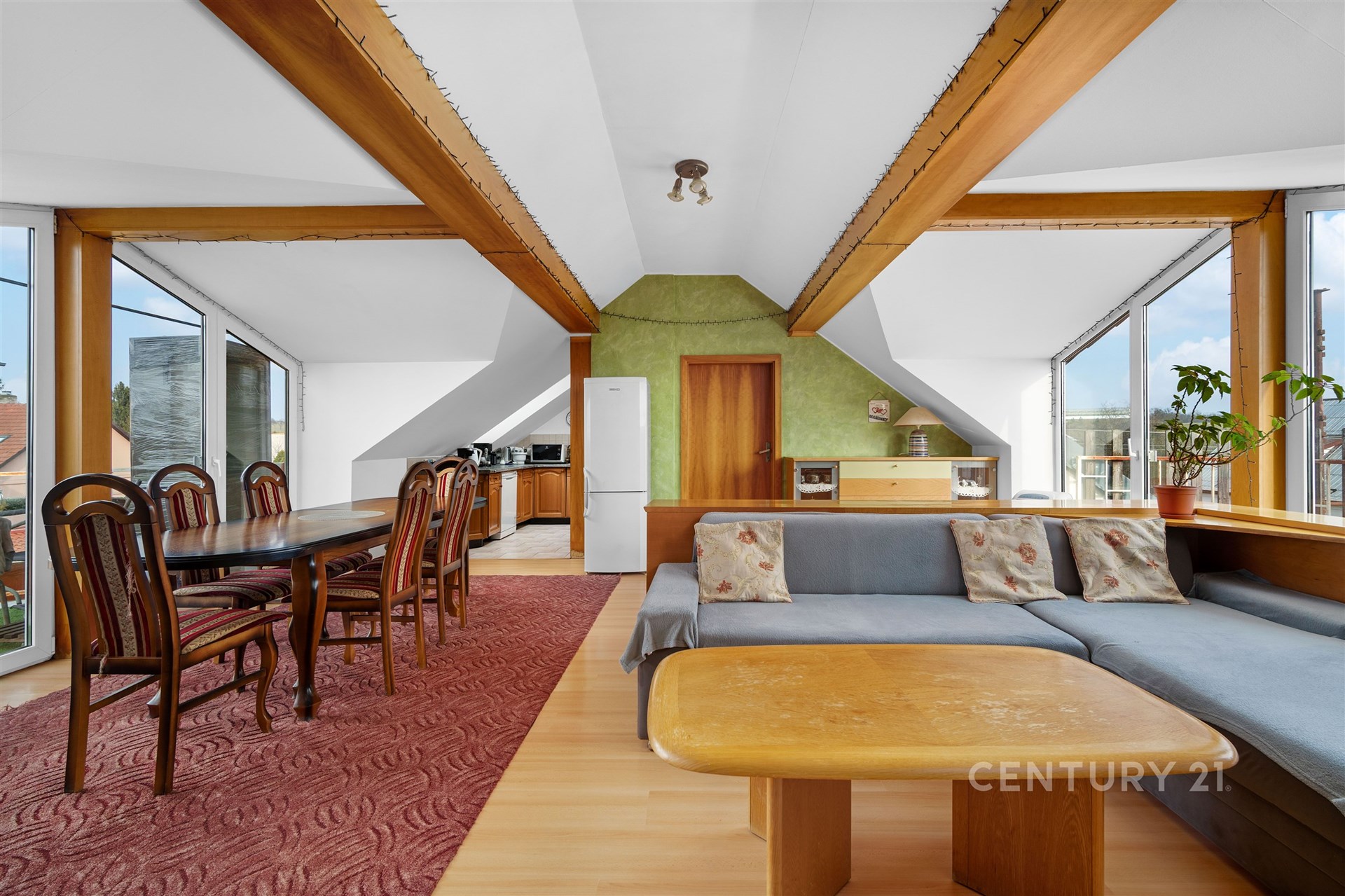 Podkrovní obývací pokoj s dřevěnými trámy, velkými okny a parketovou podlahou, spojený s jídelním koutem a moderní kuchyní.