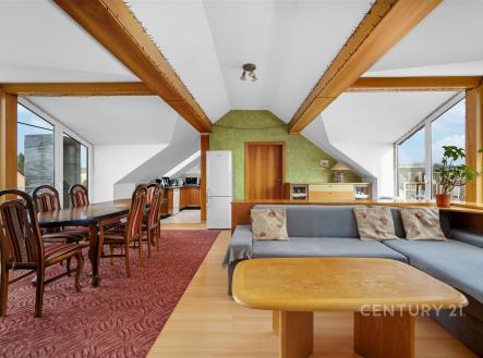 Podkrovní obývací pokoj s dřevěnými trámy, velkými okny a parketovou podlahou, spojený s jídelním koutem a moderní kuchyní. | Prodej bytu, 3+kk, 127 m²