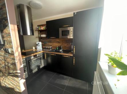 Moderní kuchyň s černými dvířky, dřevěným obložením a dlažbou. Vestavěné spotřebiče a velká okna zajišťují světlý interiér. | Prodej bytu, 2+kk, 54 m²