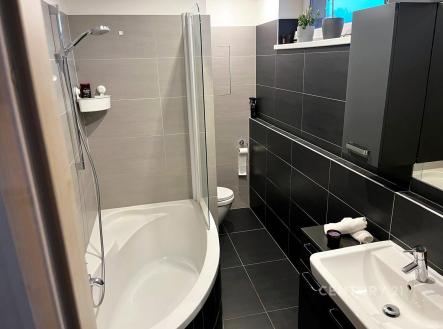 Moderní koupelna s dlažbou, rohovou vanou a tmavými obklady, nabízí světlý a elegantní design. | Prodej bytu, 2+kk, 54 m²