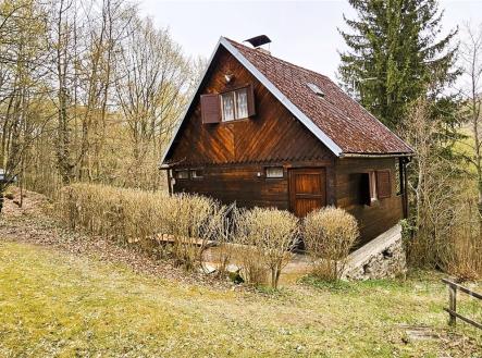 Chata v přírodě, dřevěná konstrukce s šikmou střechou, obklopena zelení a stromy, nabízí klidné prostředí a rustikální styl bydlení. | Prodej - chata/rekreační objekt, 50 m²