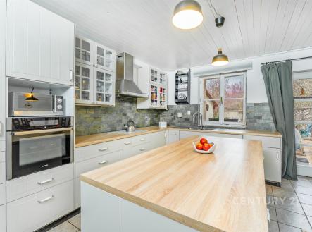 Moderní kuchyň s dřevěnou pracovní deskou, vestavěnými spotřebiči a velkými okny. Světlý prostor je doplněn dlažbou a bílými skříněmi. | Prodej - dům/vila, 217 m²