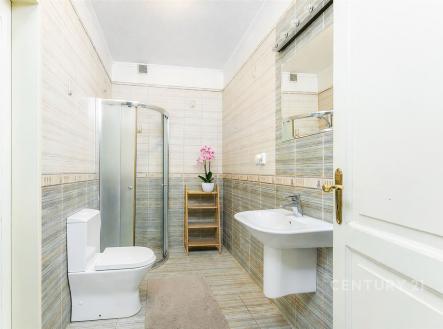 Moderní koupelna s dlažbou, sprchovým koutem a velkým zrcadlem. Světlý interiér, decentní obklady a bílá sanita vytvářejí elegantní vzhled. | Prodej - dům/vila, 217 m²