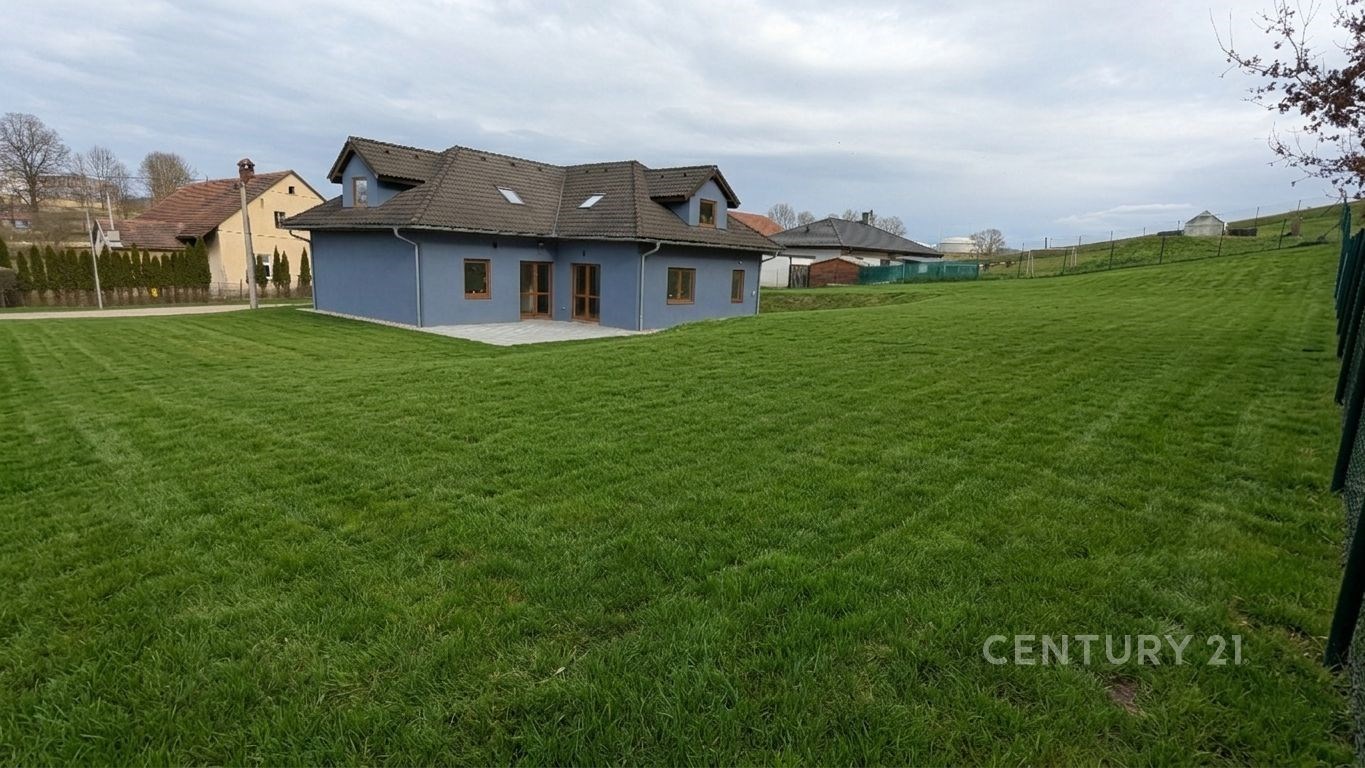 Rozlehlá zahrada s trávníkem a moderním rodinným domem v pozadí, dřevěná terasa, šikmá střecha, klidná lokalita.