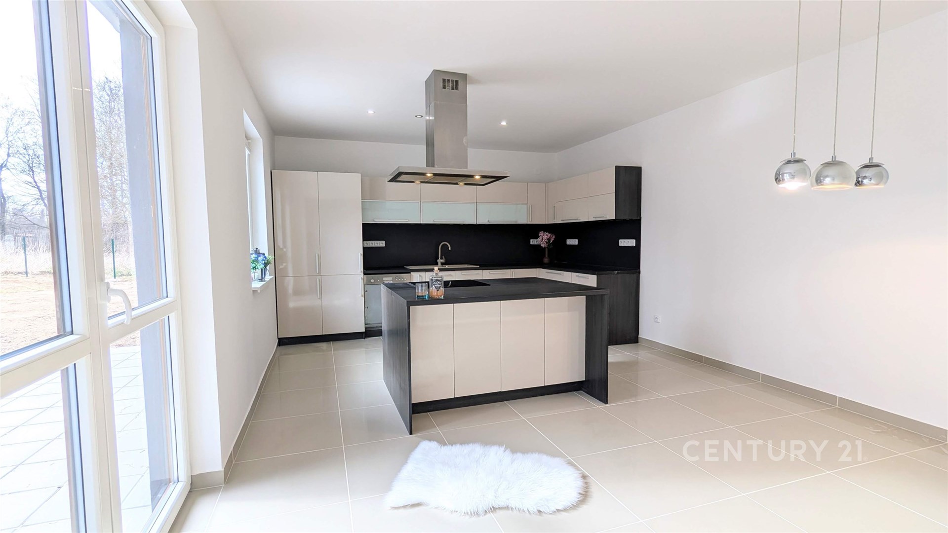 Moderní kuchyň s dlažbou, velká okna zajišťující světlost, elegantní černobílá linka s centrálním ostrůvkem, závěsná svítidla přidávají styl.
