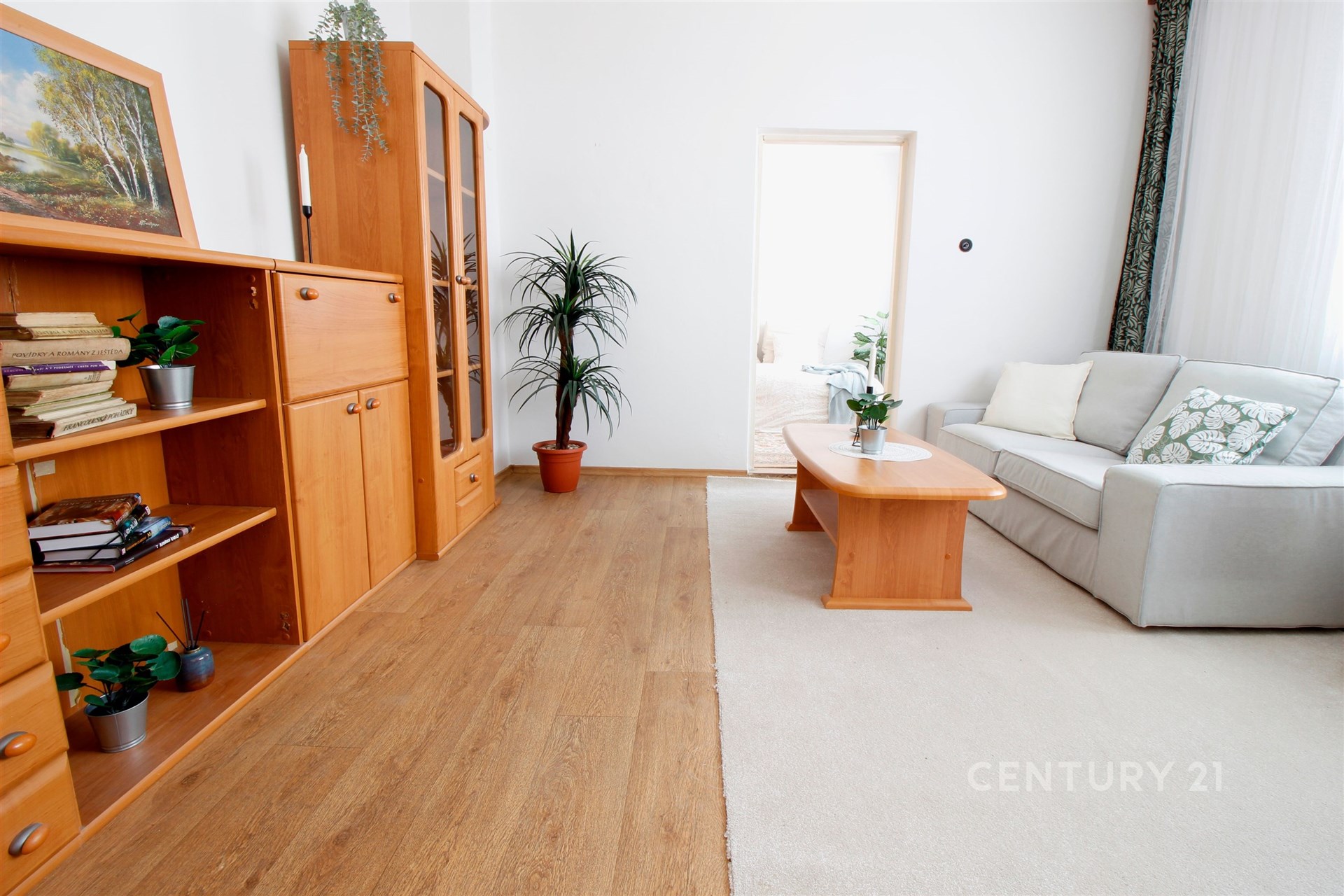 Světlý obývací pokoj s dřevěnými skříněmi, parketovou podlahou a velkými okny, doplněný moderním světlým nábytkem a zelenými rostlinami.