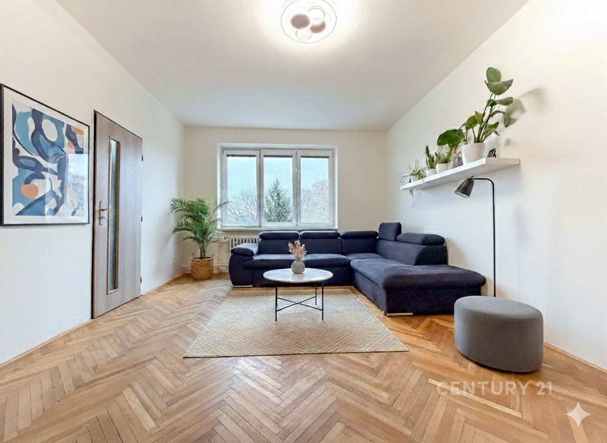 Světlý obývací pokoj s parketovou podlahou, velkými okny a moderním nábytkem, doplněný o dekorativní rostliny a minimalistický styl.