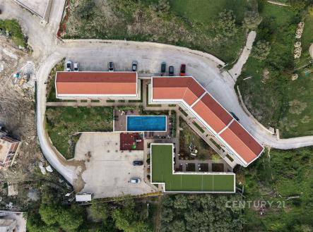Letecký pohled na moderní bytový komplex s červenou střechou a venkovním bazénem, obklopený zelení, parkovištěm a svěžími zahradami. | Prodej bytu, 1+kk, 32 m²