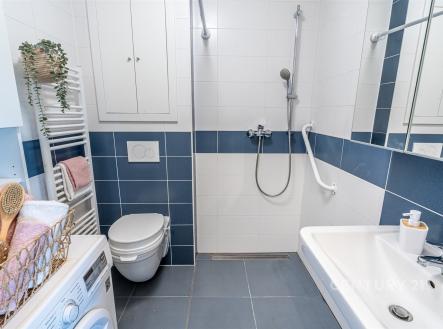 Kompaktní koupelna, moderní styl s dlažbou, sprchovým koutem a modro-bílými obklady. Obsahuje závěsnou toaletu a praktickou pračku. | Prodej bytu, 1+kk, 37 m²