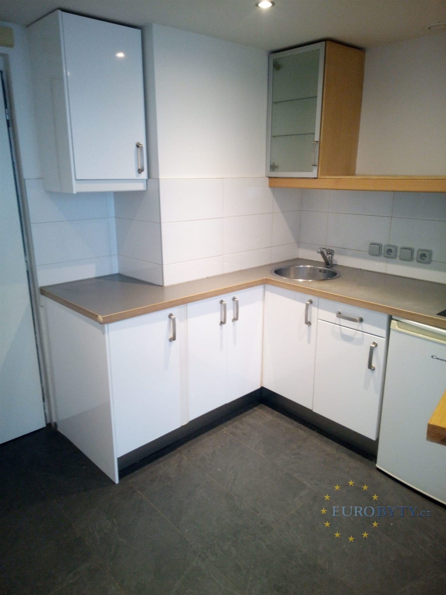 Moderní kuchyň s bílými skříňkami, dlažbou na podlaze a rohovým pracovním pultem, ideální pro malé prostory.