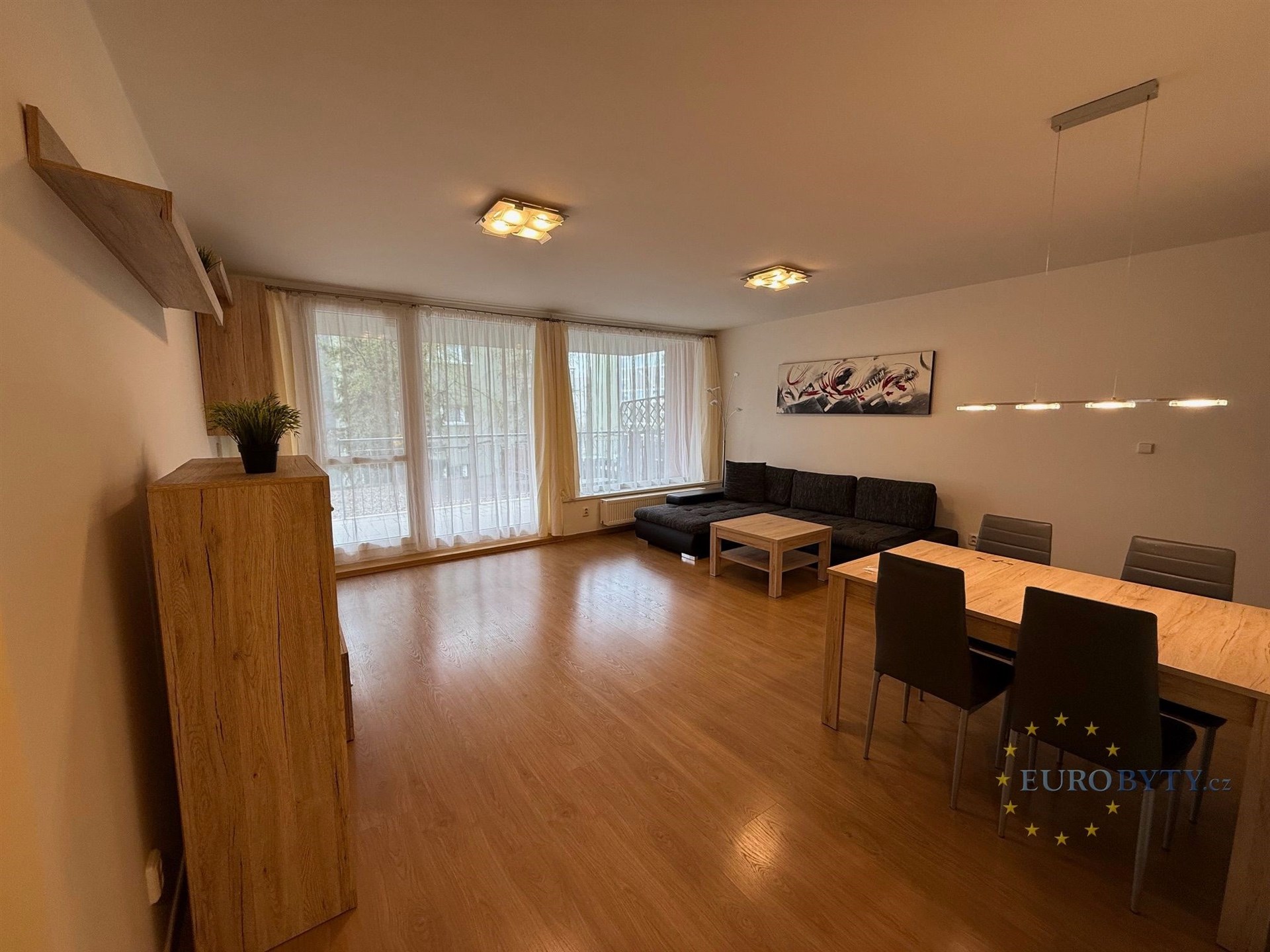Světlý obývací pokoj s parketovou podlahou, velkými okny a moderní jídelní sestavou, nabízí komfortní prostor pro rodinné posezení.
