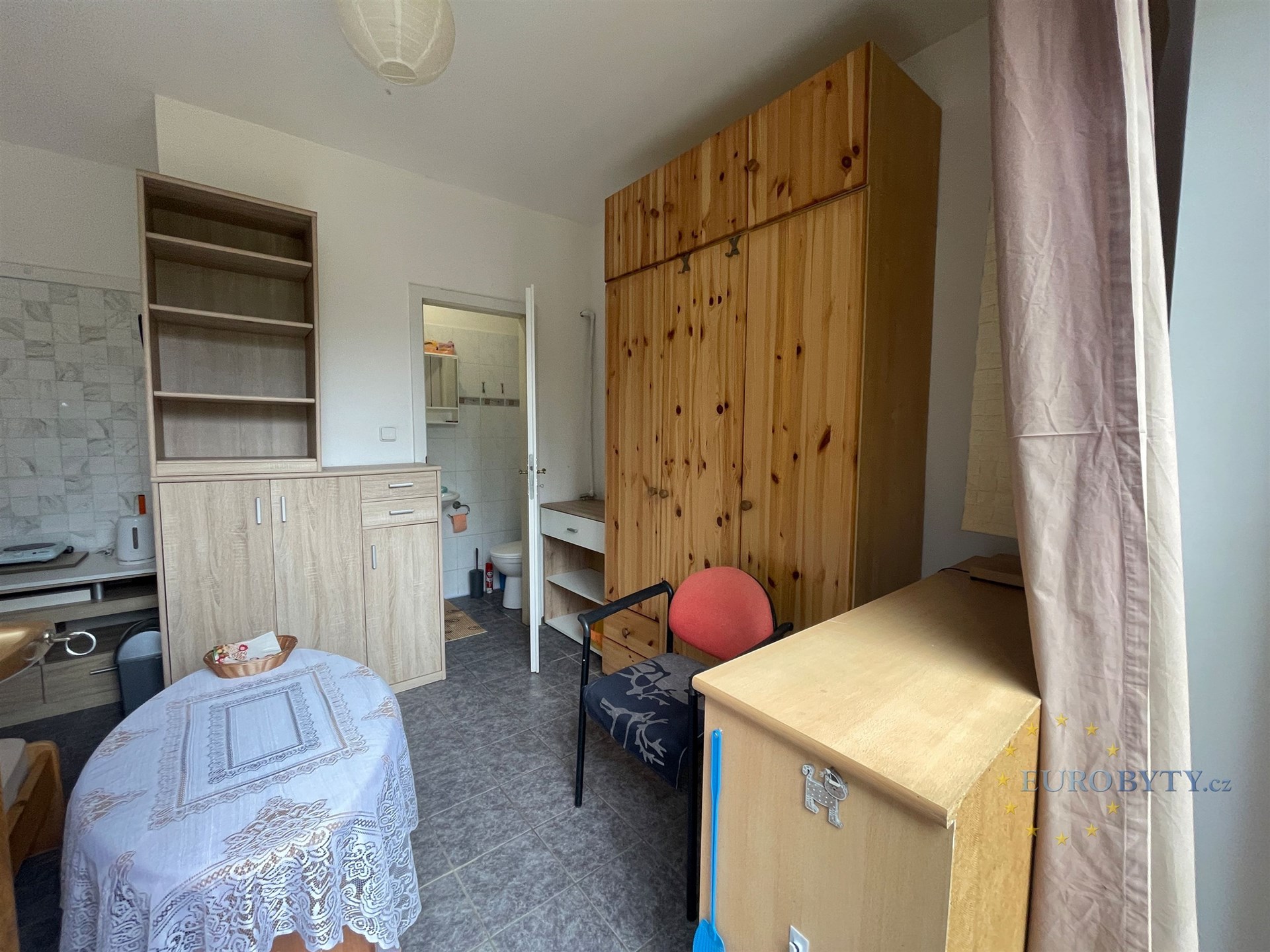 Kompaktní pokoj s dlažbou, vestavěnými skříněmi a dřevěným nábytkem, vedle moderní kuchyňky a přístupem do koupelny, ideální pro single bydlení.