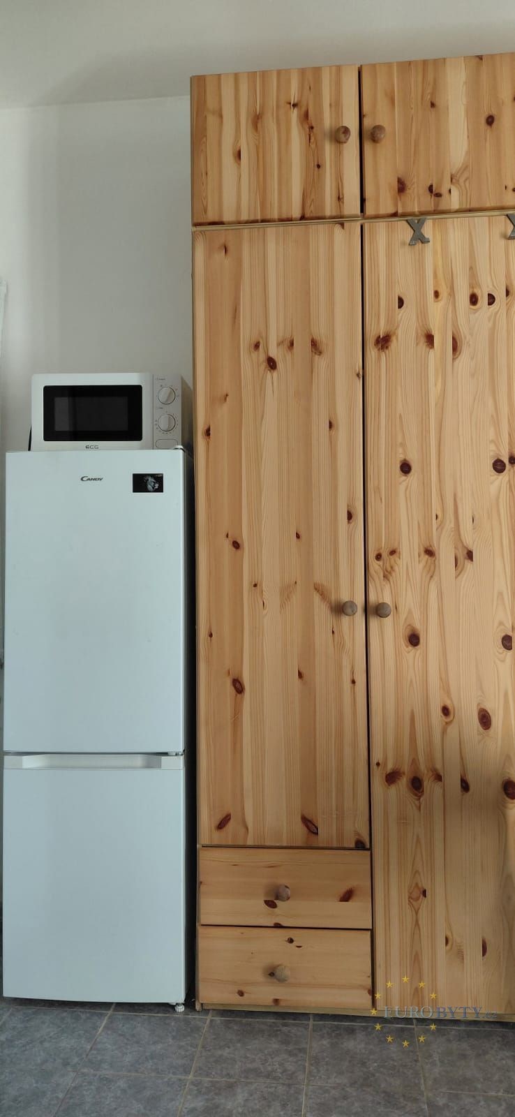 Kuchyň s lednicí, mikrovlnnou troubou a dřevěnými vestavěnými skříněmi, moderní vzhled a dlažba na podlaze pro snadnou údržbu.