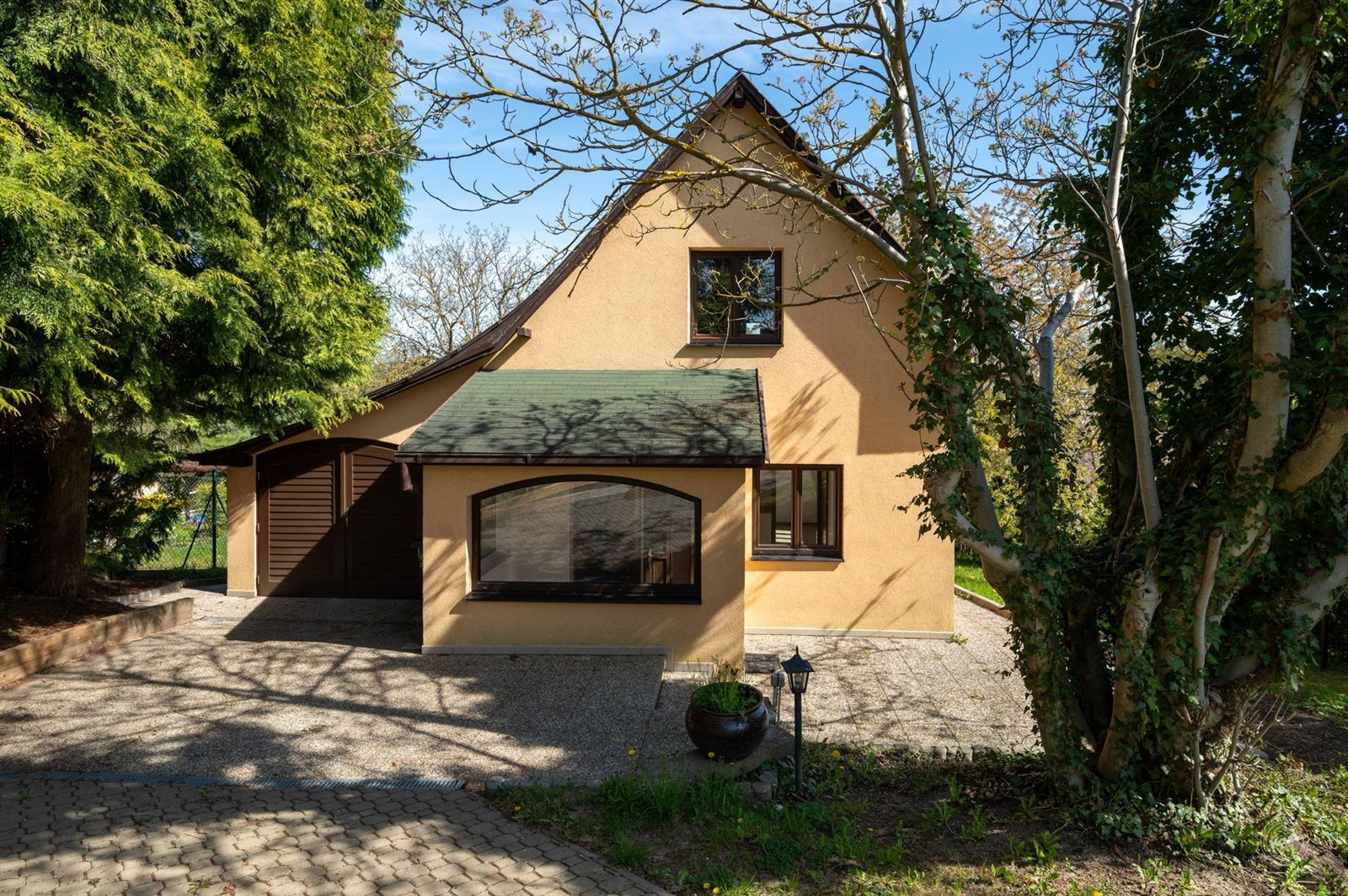 Rodinný dům s moderní fasádou, obklopený zelení. Velká okna poskytují dostatek světla, pěkný udržovaný pozemek, klidná lokalita.