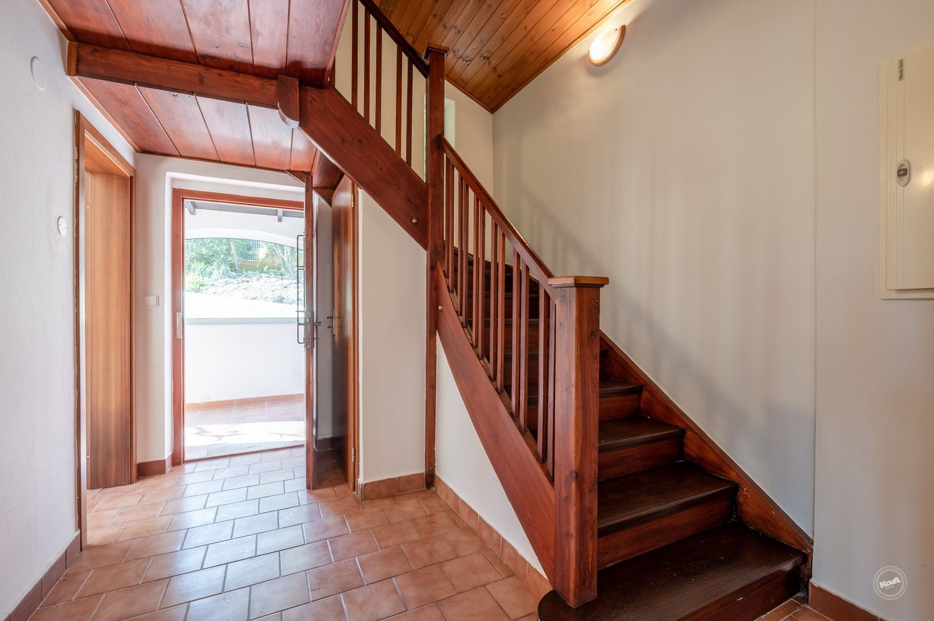 Chodba s dřevěným schodištěm, dlažba na podlaze, světlé stěny. Vstup do domu s přírodními materiály, prostorný a dobře osvětlený prostor.