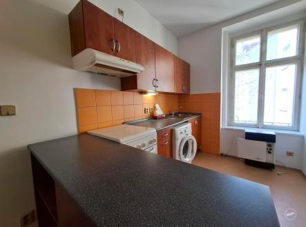 Kompaktní kuchyň s oranžovým obkladem, vestavěnými skříněmi a laminátovou pracovní deskou, světlá díky velkému oknu. | Pronájem bytu, 1+1, 31 m²