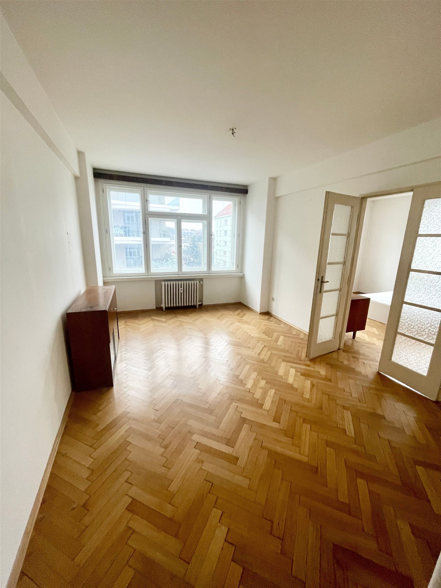 Prostorný obývací pokoj s parketovou podlahou, velkými okny a bílými stěnami, ideální pro moderní bydlení.