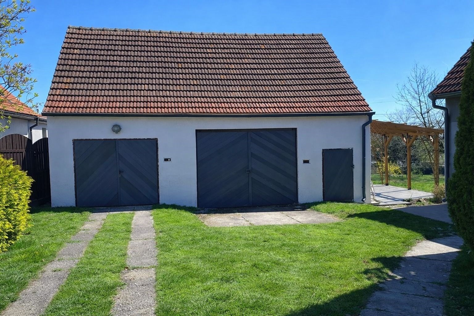 Praktická garáž s dvojitými vraty, příjezdová cesta s dlážděným povrchem, vedlejší prostor pro zastřešené parkování, dobře udržovaná zahrada.