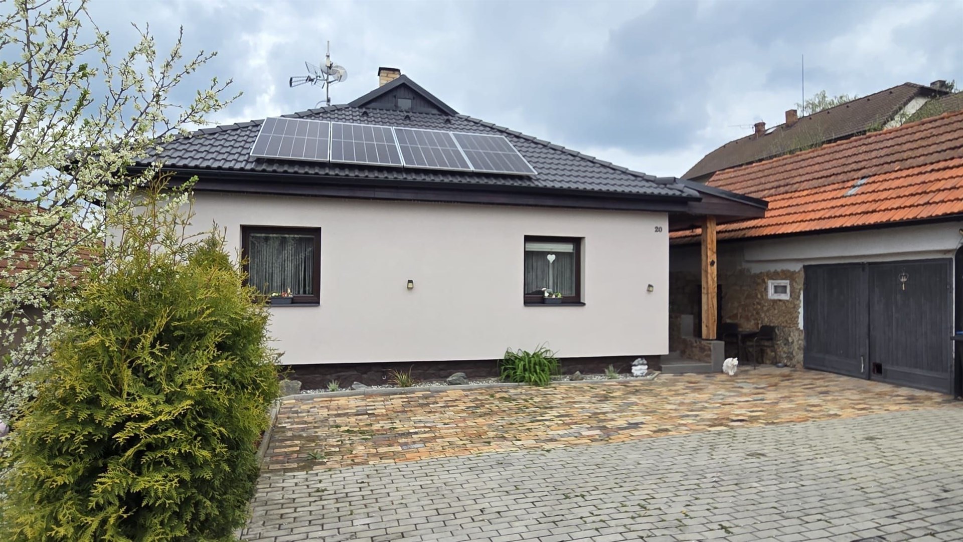 Fasáda rodinného domu s moderními solárními panely, zámkovou dlažbou a dřevěnou pergolou na kryté parkování, poklidná rezidenční čtvrť.