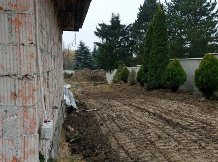 Zahrada se stromky a cihlovou zdí, vedle nezastavěného pozemku. Viditelné surové cihly domu. | Prodej - dům/vila, 925 m²