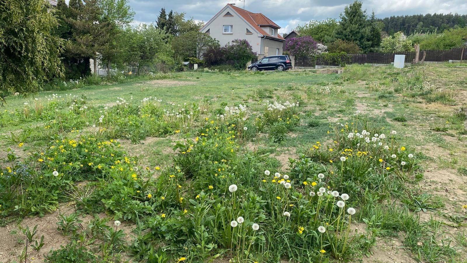 Stavební pozemek s rovinatým terénem obklopený zelení a květinami, ideální pro stavbu rodinného domu, klidná rezidenční lokalita.