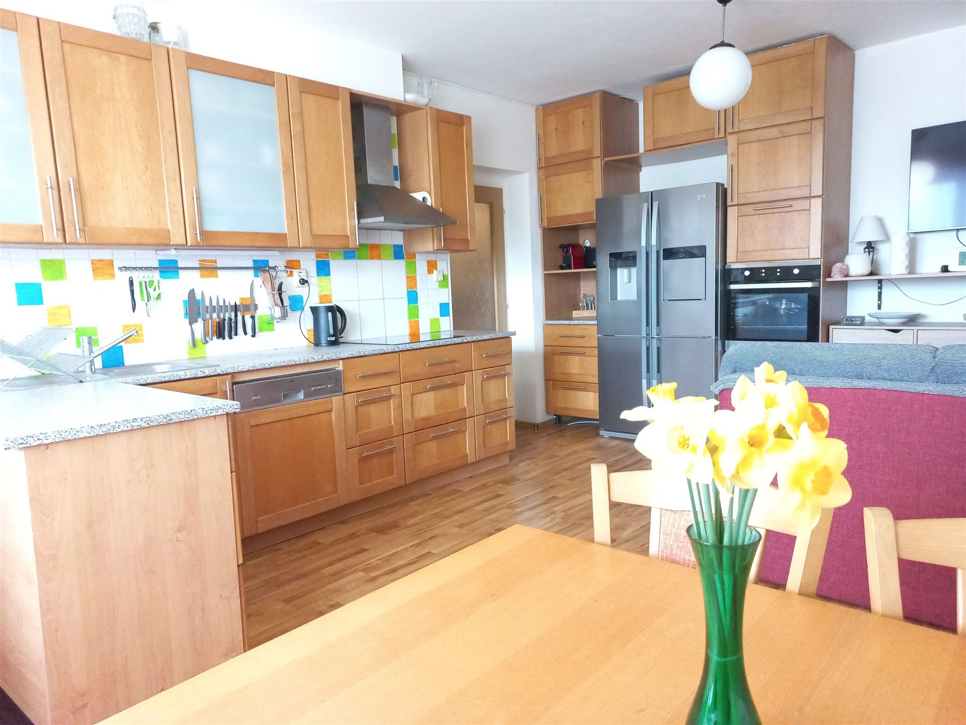 Kuchyň s dřevěnými skříňkami a parketovou podlahou, moderní spotřebiče a barevný obklad, prostorný a světlý interiér.