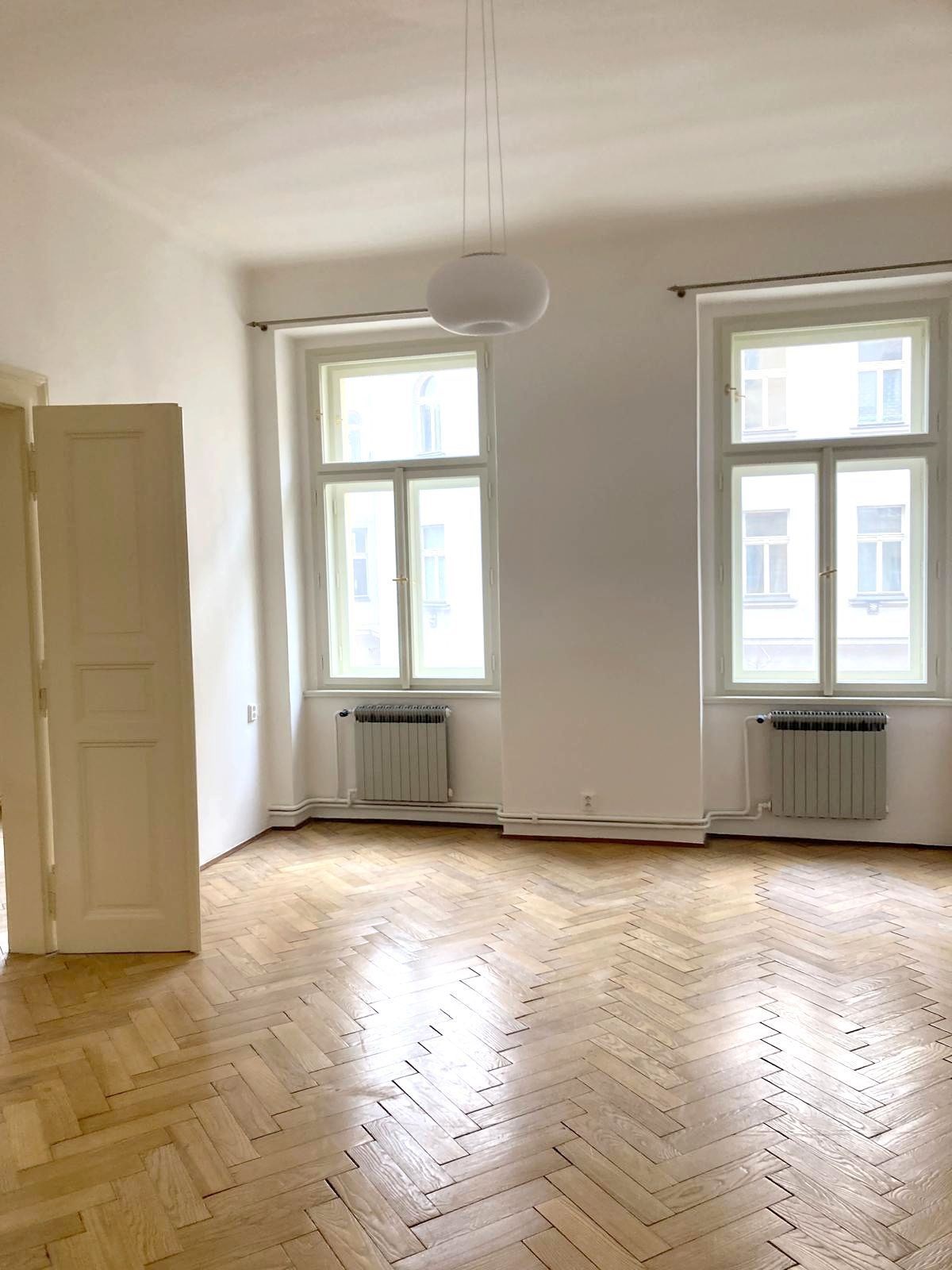 Světlý obývací pokoj s parketovou podlahou, velká okna poskytující přirozené světlo, v dobrém stavu připravený k nastěhování.