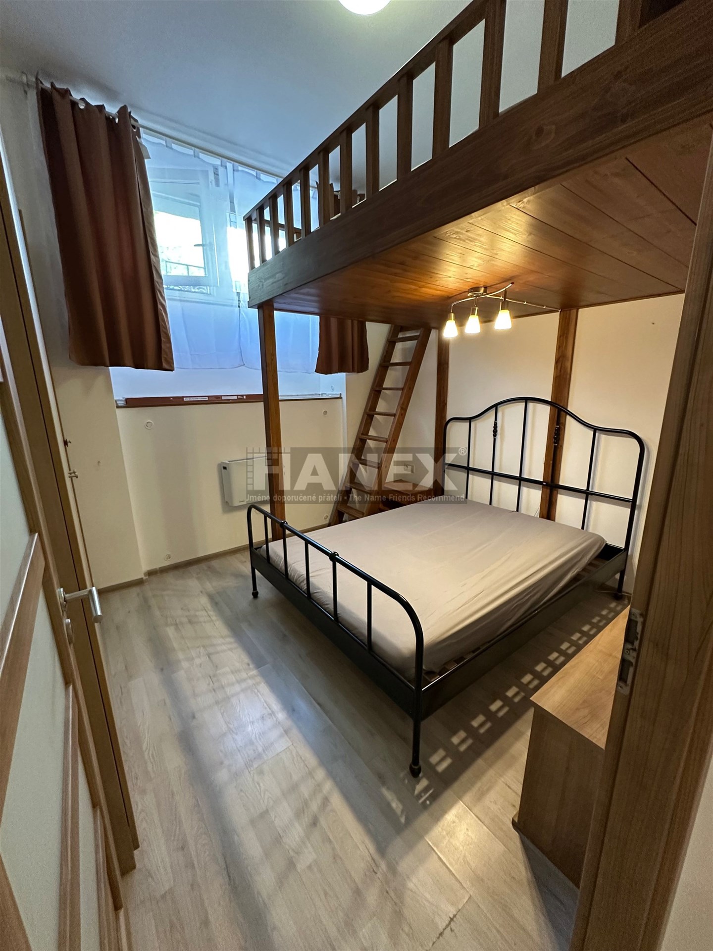 Malá ložnice s dřevěným patrem, kovovou postelí a parketovou podlahou. Světlé provedení, ideální pro úsporu místa.