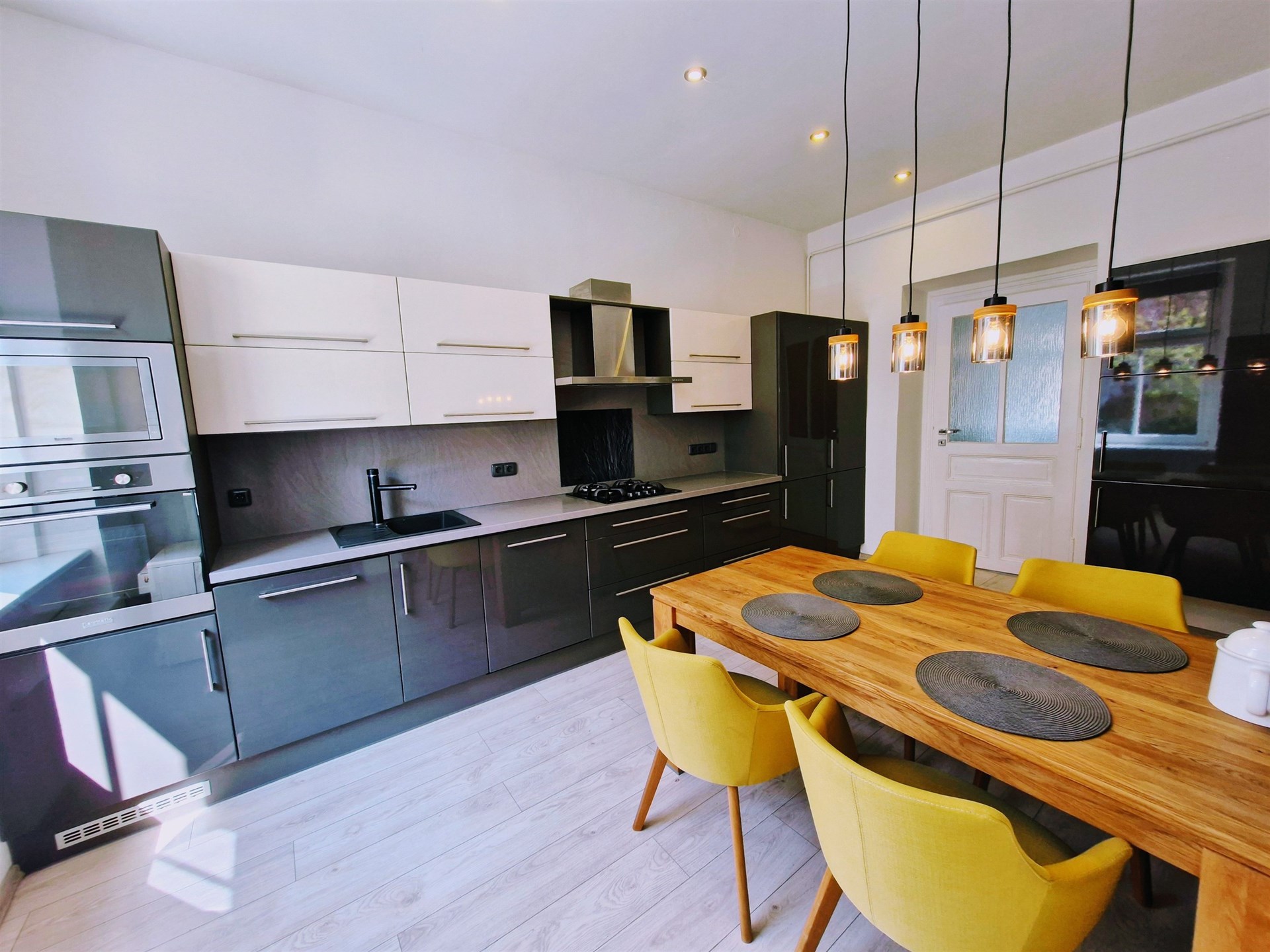 Moderní kuchyň s parketovou podlahou, vestavěné spotřebiče, dřevěný jídelní stůl, světlý interiér, stylové osvětlení.