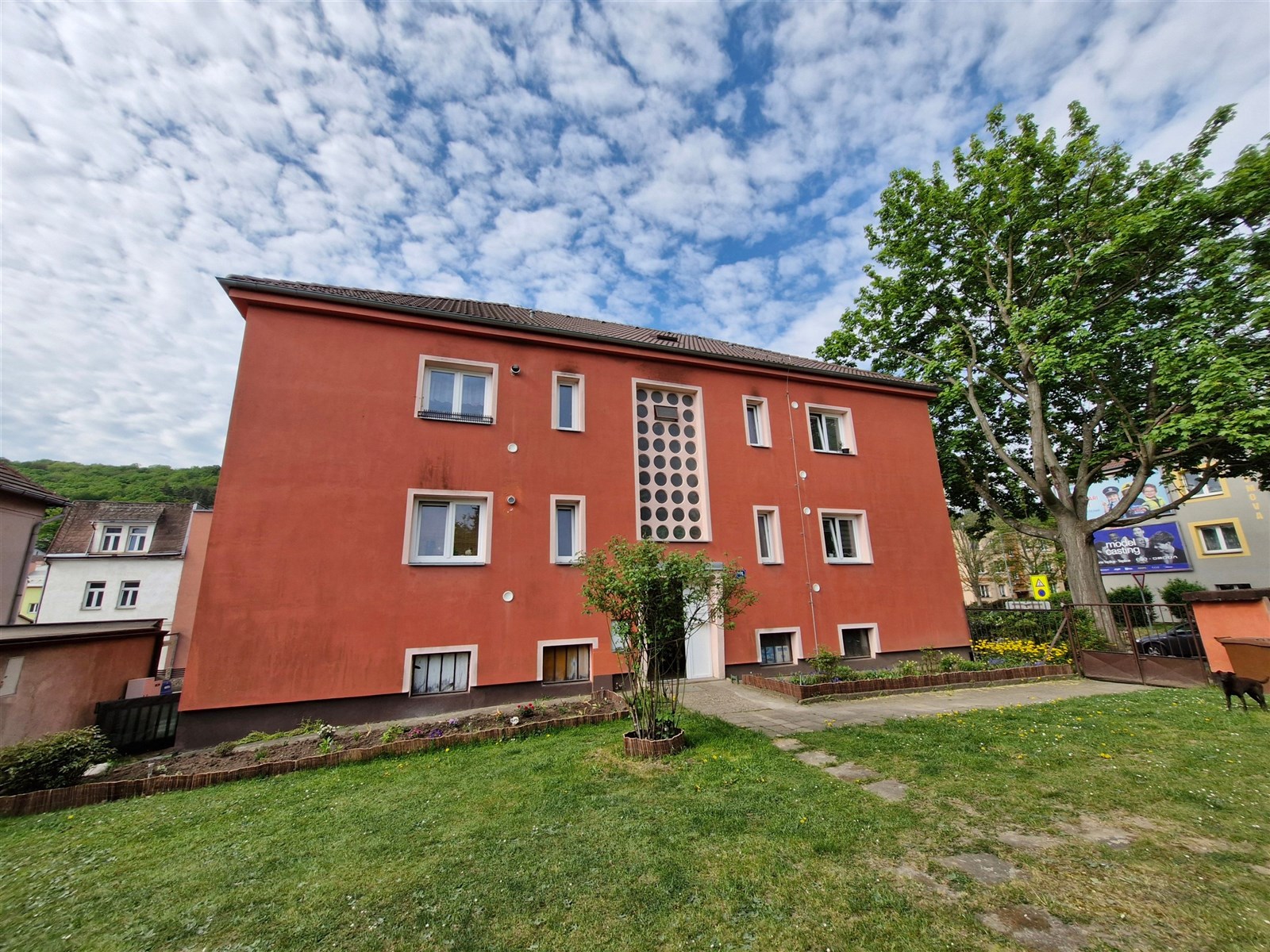 Červený bytový dům s moderní fasádou a velkými okny, obklopený upravenou zahradou, v klidné rezidenční lokalitě.