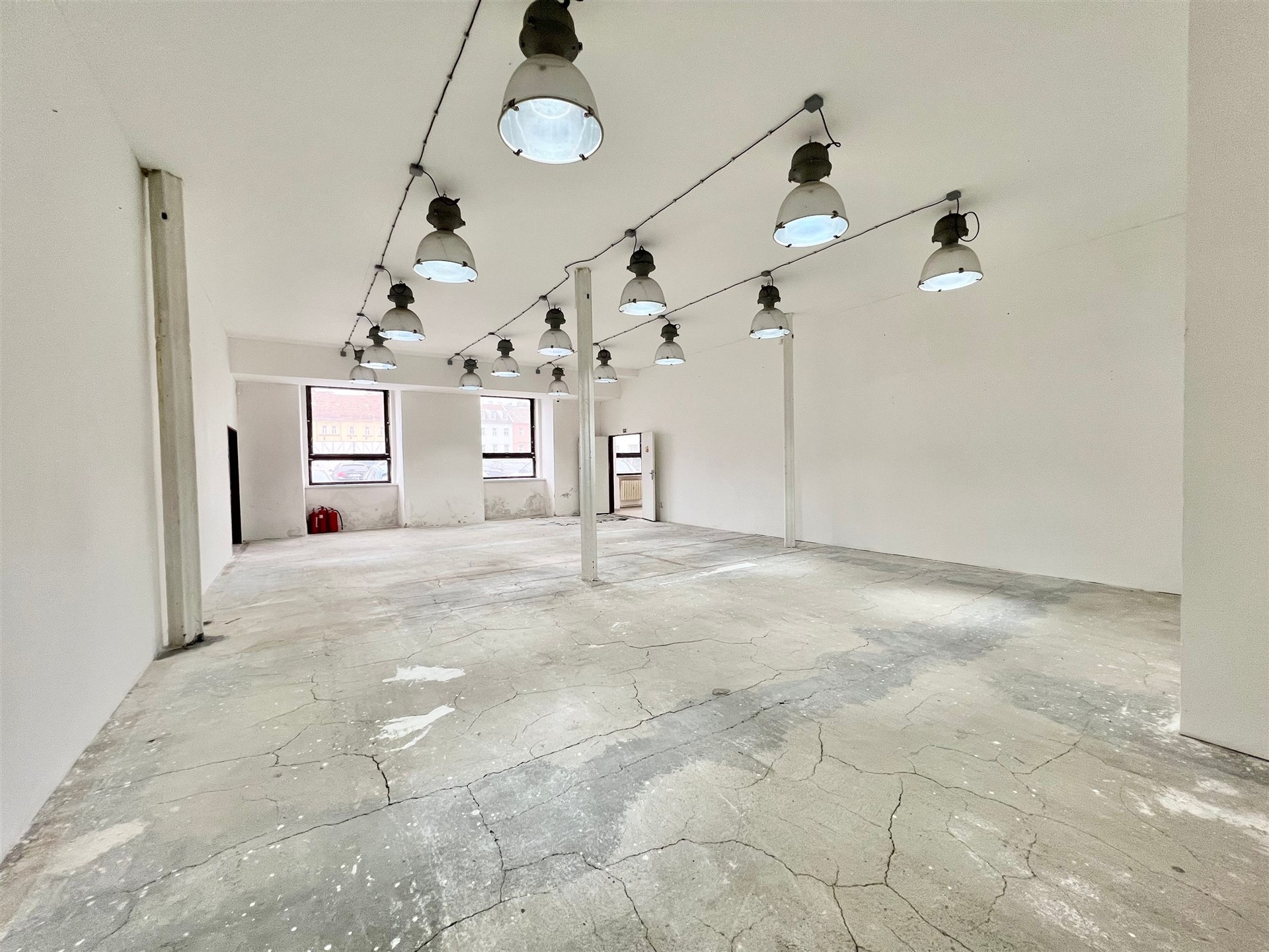 Průmyslový prostor s betonovou podlahou, velkými okny a stropními svítidly, vhodný pro kreativní využití nebo rekonstrukci