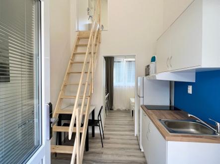 Kompaktní kuchyň s dřevěnou podlahou, moderními bílými skříňkami a schodištěm na patro, umožňuje efektivní využití prostoru. | Pronájem bytu, 2+1, 35 m²