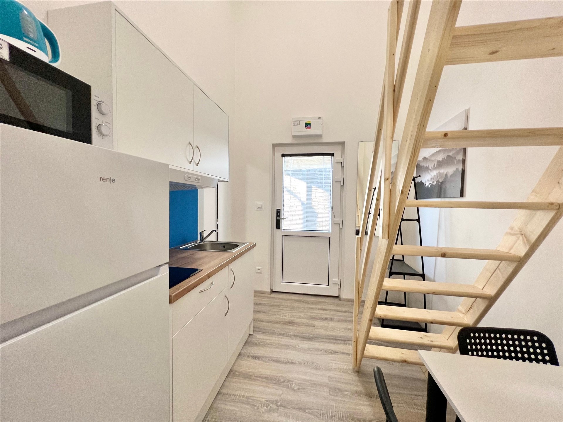 Malá kuchyň s moderními spotřebiči, dřevěným schodištěm a světlou vinylovou podlahou, ideální pro kompaktní bydlení.