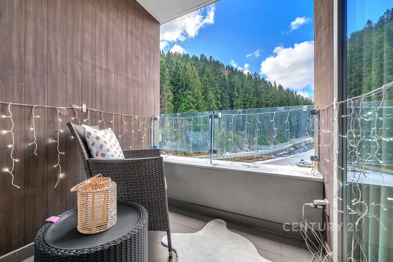 Balkón s moderní dlažbou a proskleným zábradlím nabízí panoramatický výhled na lesní krajinu, útulný kout s ratanovým nábytkem a světelným řetězem.