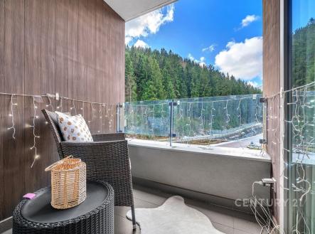 Balkón s moderní dlažbou a proskleným zábradlím nabízí panoramatický výhled na lesní krajinu, útulný kout s ratanovým nábytkem a světelným řetězem. | Prodej bytu, 2+kk, 58 m²