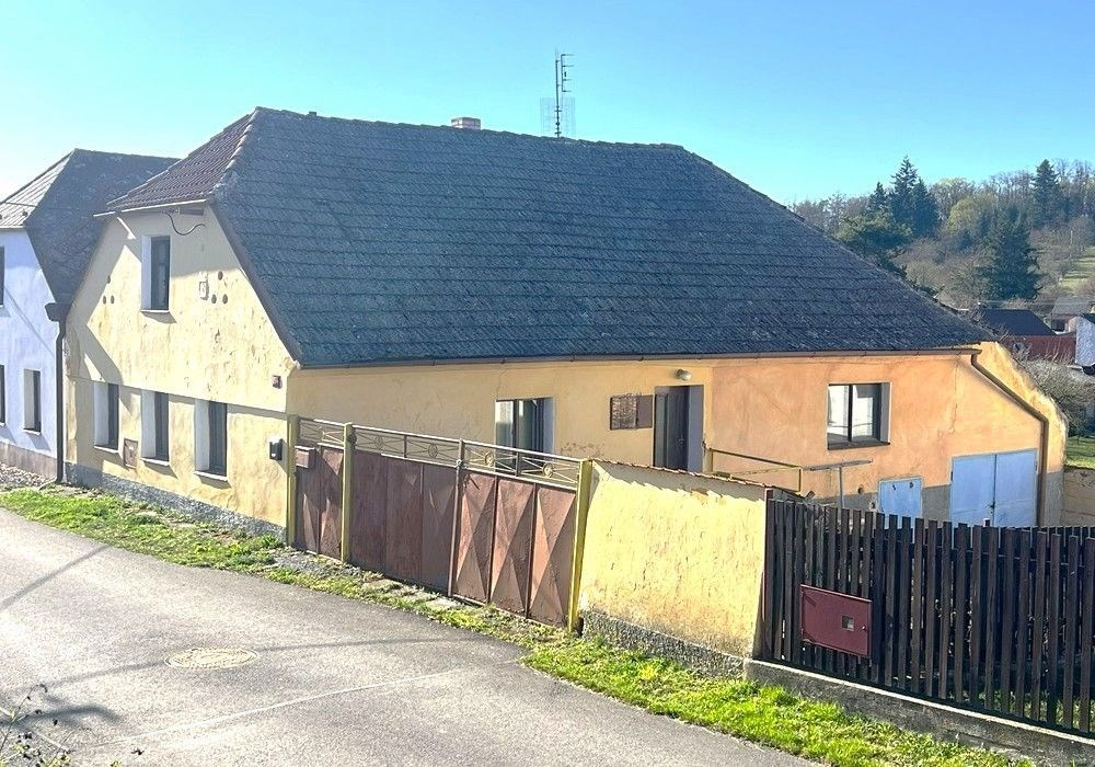 Kamenný venkovský dům s tradiční šikmou střechou a dřevěnými okenicemi se nachází na klidné ulici v malebné krajině.