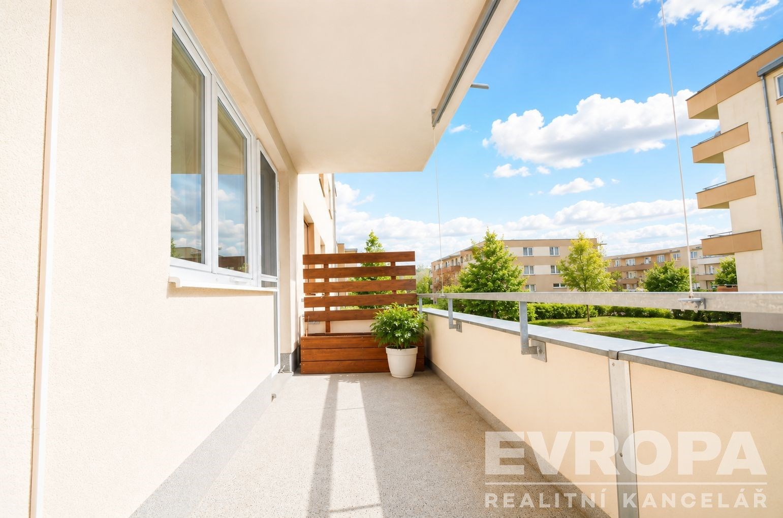 Prostorný balkon s moderní dlažbou, velká okna propouštějící mnoho světla, výhled na zelené okolí, světlý a udržovaný stav.