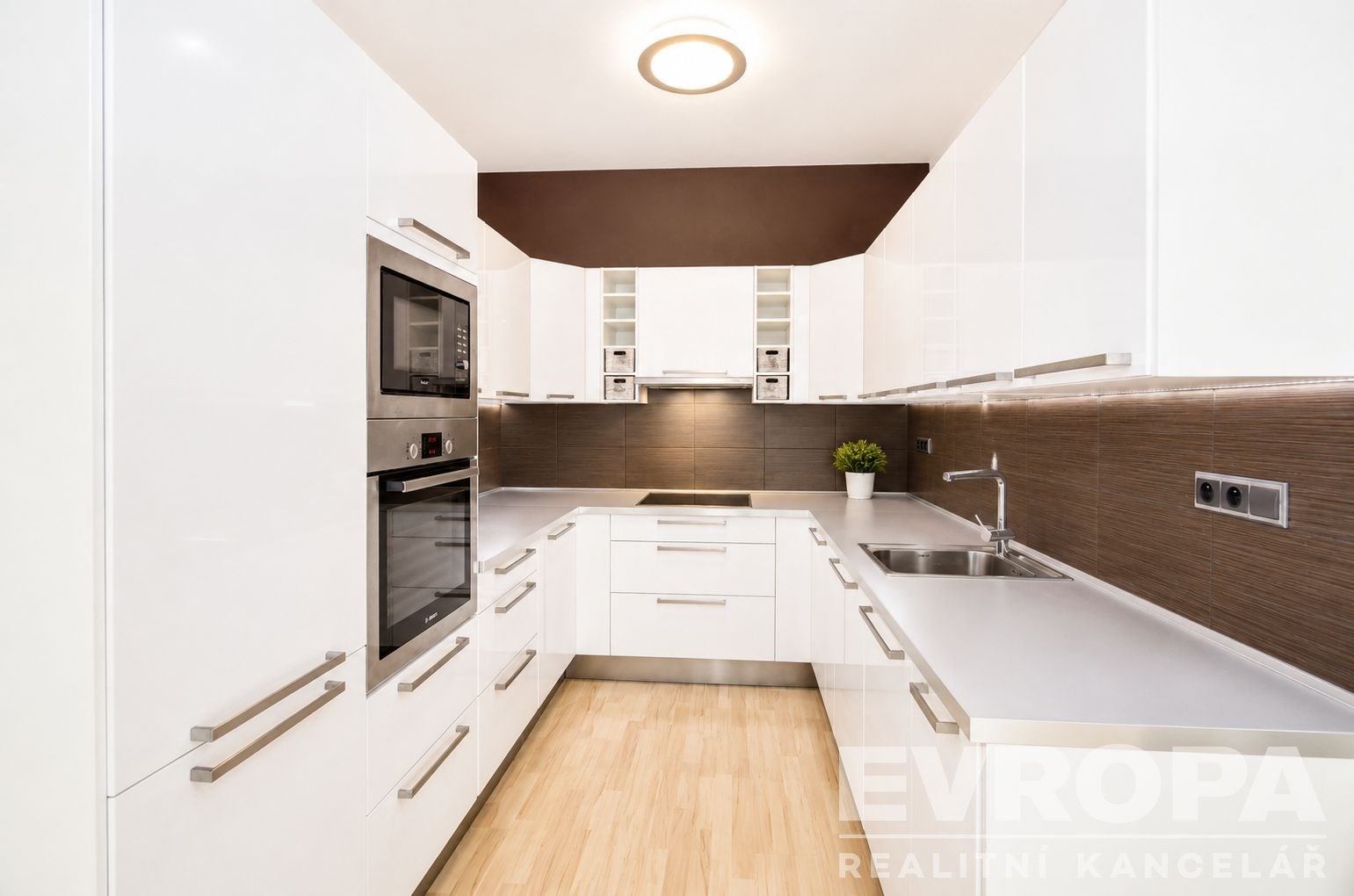 Moderní kuchyň s bílými skříňkami, vestavěnými spotřebiči a parketovou podlahou. Světlý prostor s moderním osvětlením a elegantním designem.