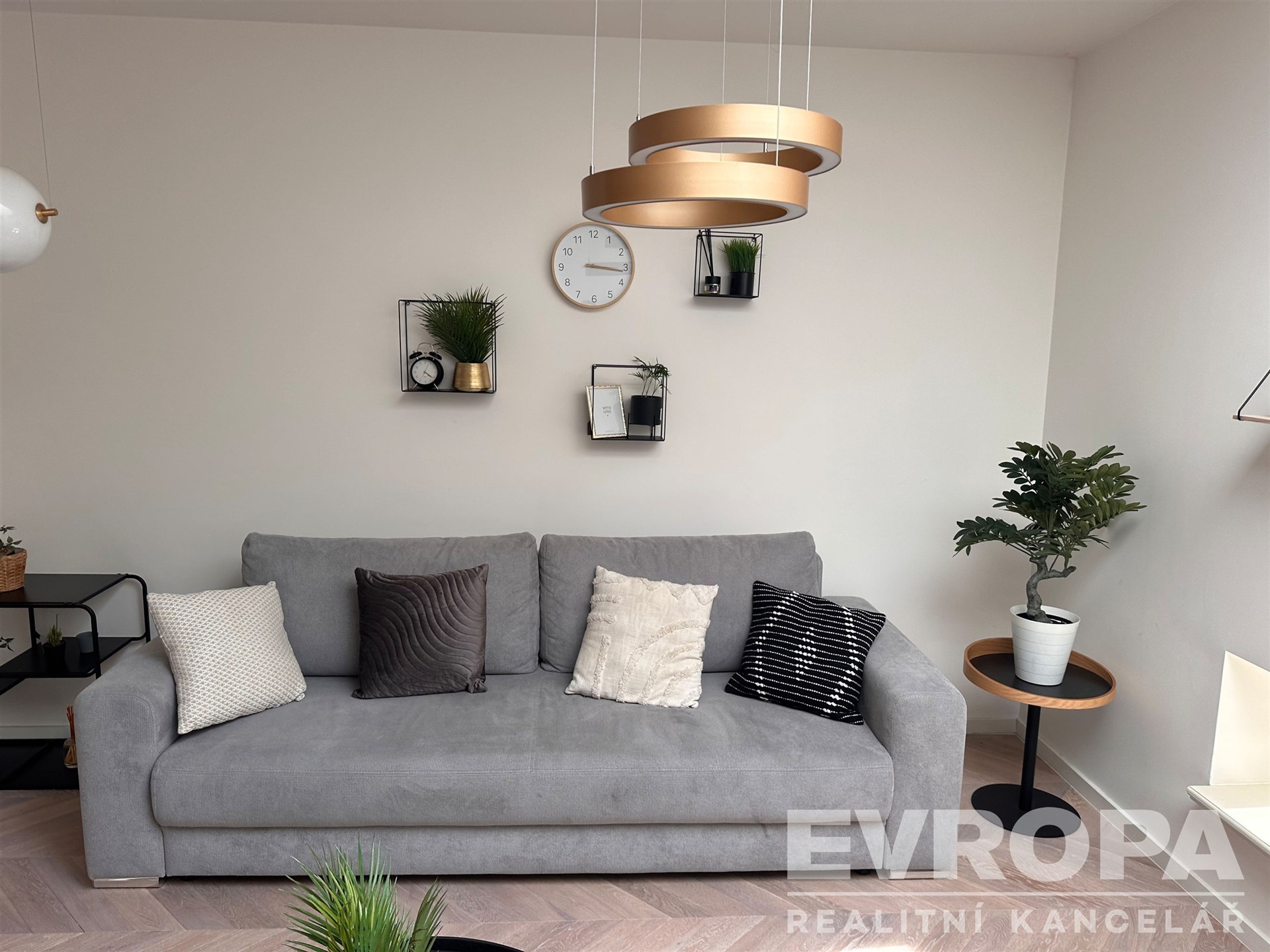 Světlý obývací pokoj s parketovou podlahou, moderní pohovkou a stylovým osvětlením, doplněn dekorativními prvky a rostlinami.