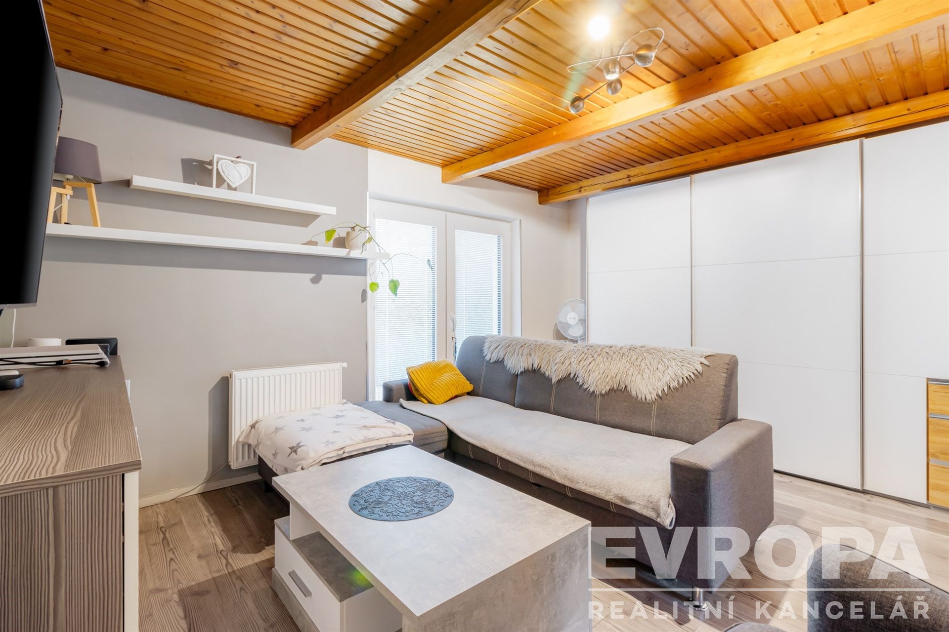 Světlý obývací pokoj s dřevěnými stropy, moderní vestavěné skříně, parketová podlaha, útulné vybavení, a elegantní nábytek.