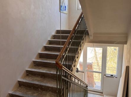 Schodiště s kovovým zábradlím, betonové stupně, světlé stěny a velká okna poskytující přirozené světlo, ideální pro modernizaci nebo rekonstrukci. | Prodej bytu, 1+kk, 27 m²