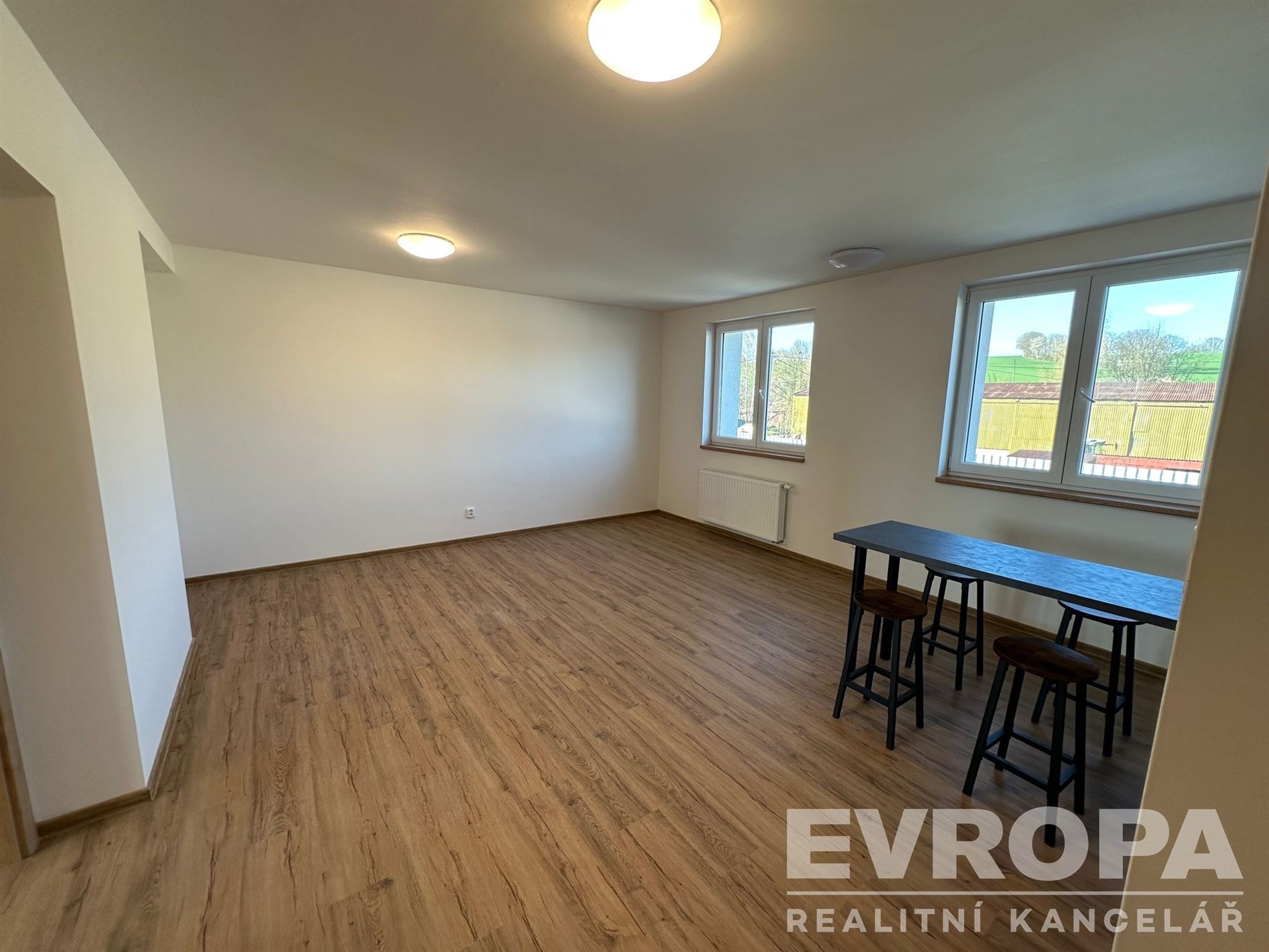 Světlý obývací pokoj s moderní parketovou podlahou a velkými okny, které poskytují dostatek přirozeného světla.