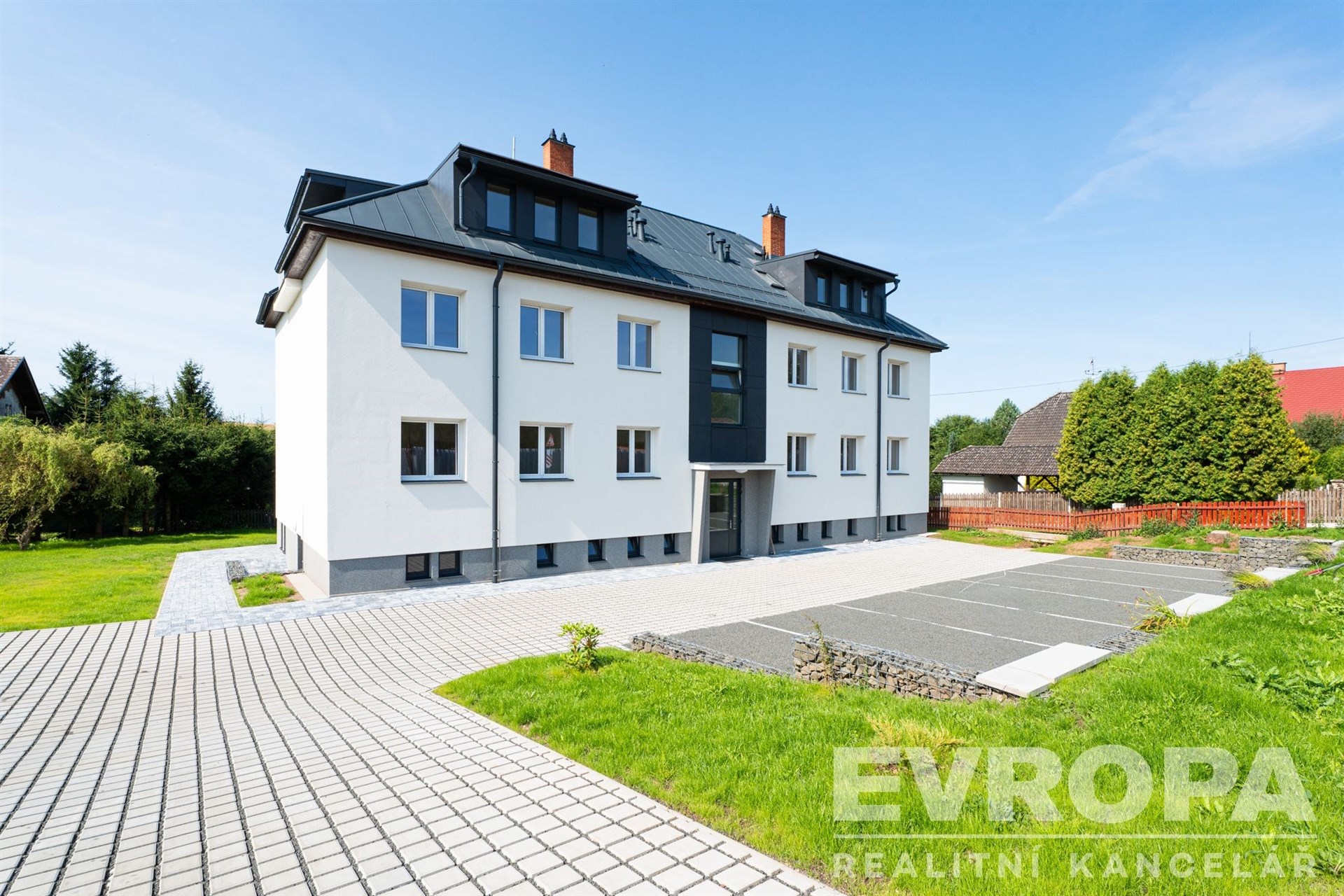 Praktický exteriér bytového domu s moderní fasádou, upravenou zahradou a dlážděným parkovacím prostorem, ideální pro rodinné bydlení.