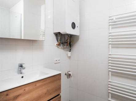 Moderní koupelna s dlažbou, dřevěnou skříňkou pod umyvadlem a bílými obklady. Obsahuje topný žebřík a novou plynovou karmu. | Pronájem bytu, 1+kk, 44 m²