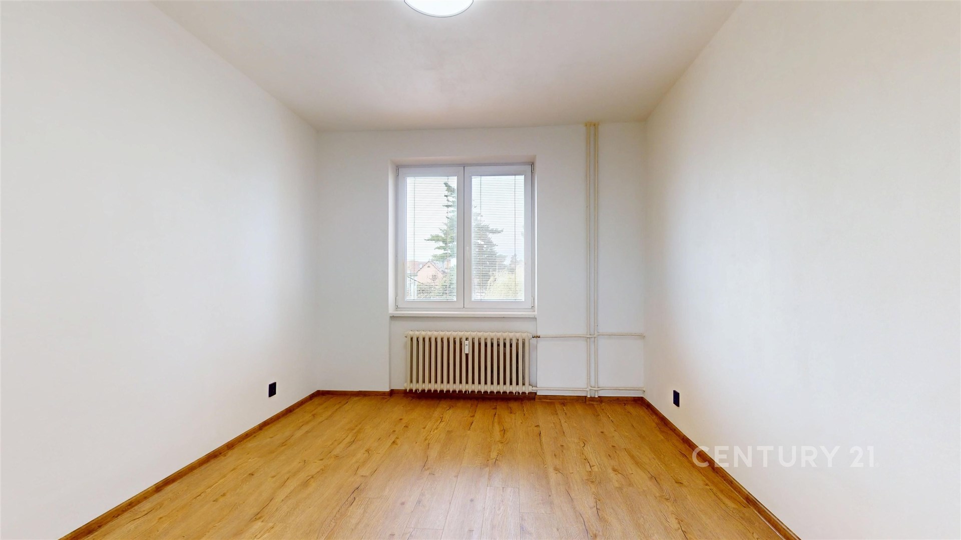 Prázdný pokoj s parketovou podlahou, světlými stěnami a velkým oknem. Radiátor pod oknem poskytuje vytápění.