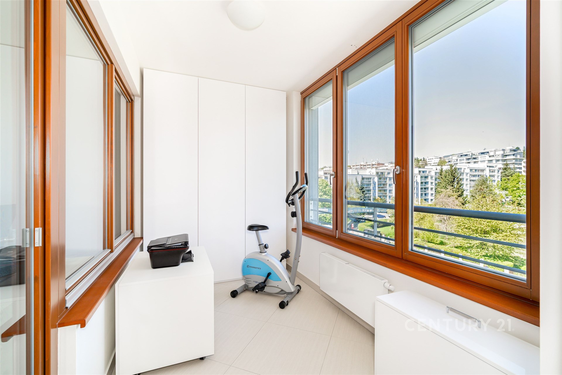 Světlá lodžie s velkými okny a dlažbou nabízí výhled na městskou krajinu, moderní design podtrhují vestavěné skříně a čisté linie.