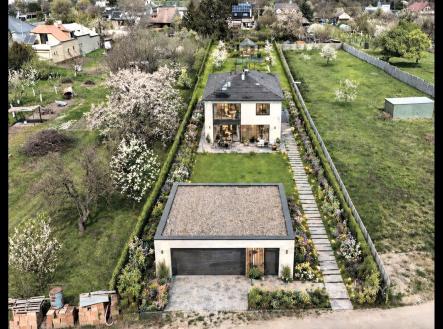 Zahrada s moderním rodinným domem a garáží, obklopená pečlivě udržovaným trávníkem a květinovými záhony, s přístupovou cestou z dlažby. | Prodej - pozemek pro bydlení, 1 057 m²